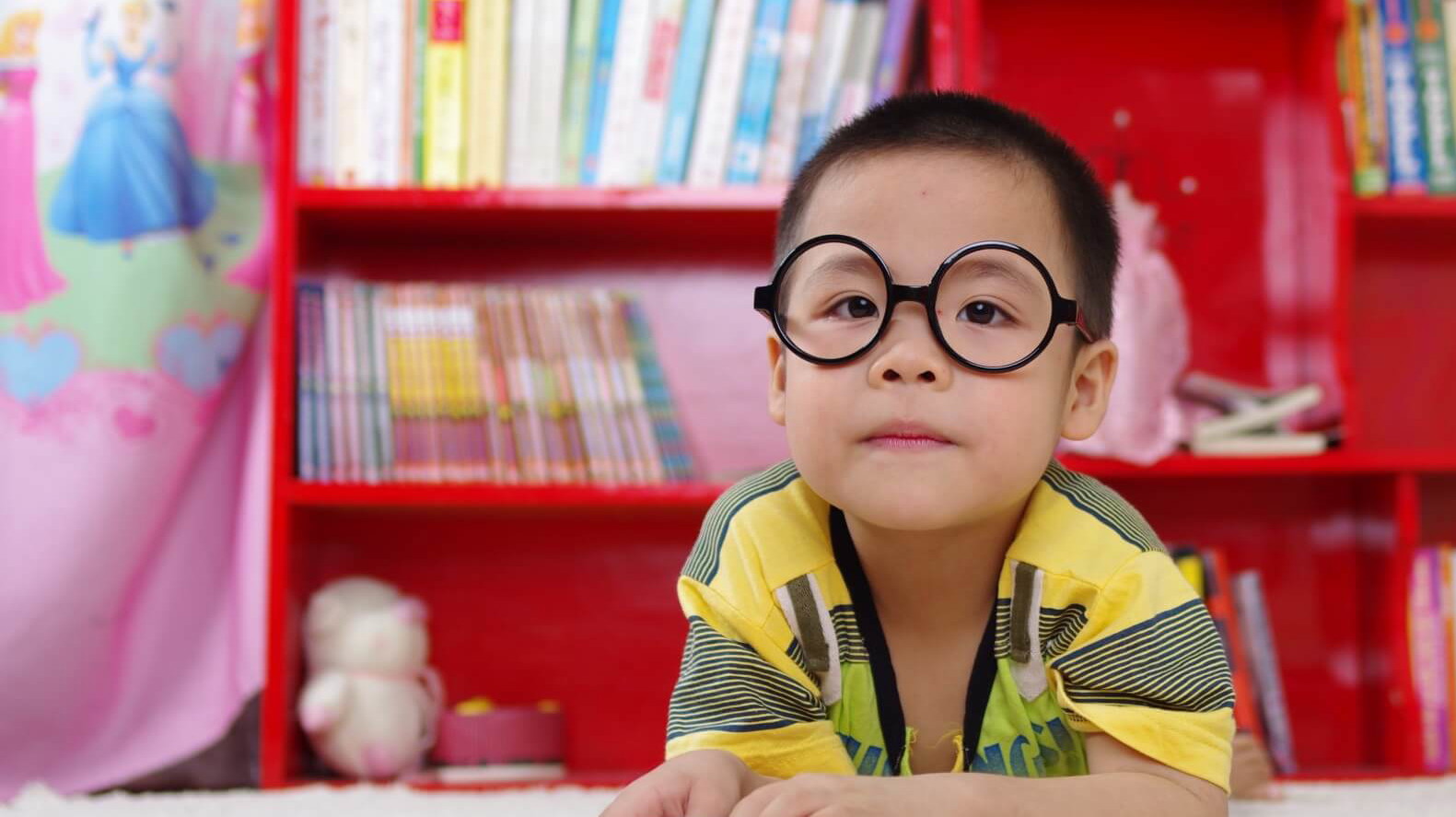 Boy with glasses reading a book sitting in front of a red bookshelf